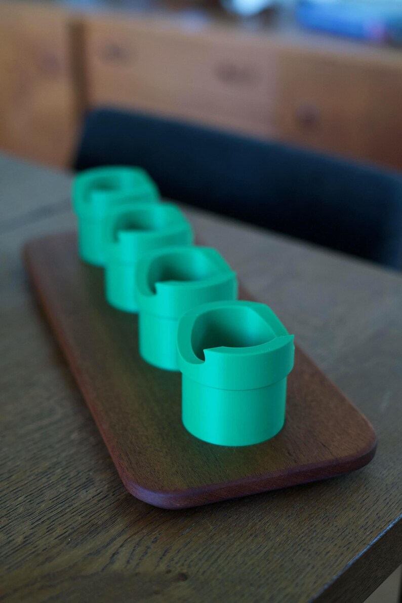 May include: Four bright green cylindrical holders with a distinct notched rim design, arranged in a single row on a rectangular wooden base. Each holder features a smooth, matte finish and a consistent shape, creating a uniform appearance.