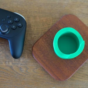 May include: A black wireless video game controller with dual analog sticks, a directional pad, and four action buttons, positioned next to a bright green cylindrical holder mounted on a rectangular, polished dark brown wooden base.
