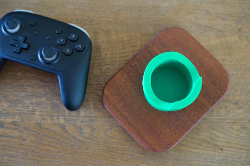 May include: A black wireless video game controller with dual analog sticks, a directional pad, and four action buttons, positioned next to a bright green cylindrical holder mounted on a rectangular, polished dark brown wooden base.