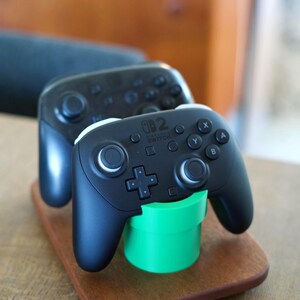 May include: Two black Nintendo Switch Pro controllers resting on a wooden base. The front controller is supported by a bright green cylindrical pipe-shaped stand. Each controller features a directional pad, two analog sticks, and buttons labeled A, B, X, and Y.