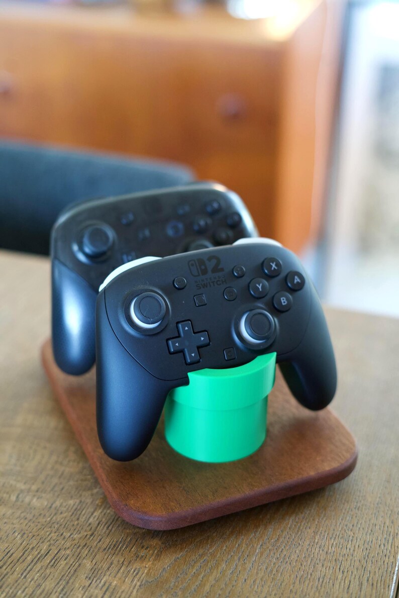 May include: Two black Nintendo Switch Pro controllers resting on a wooden base. The front controller is supported by a bright green cylindrical pipe-shaped stand. Each controller features a directional pad, two analog sticks, and buttons labeled A, B, X, and Y.