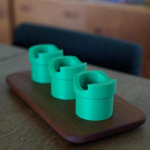 May include: Three bright green cylindrical holders with a distinct notched rim design. Each holder features a smooth, matte finish and a hollow center, arranged in a linear row on a rectangular wooden base with rounded edges and a dark brown grain.
