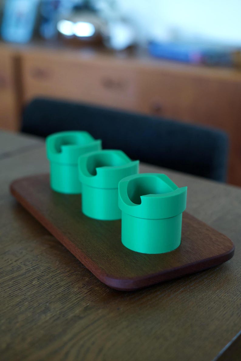 May include: Three bright green cylindrical holders with a distinct notched rim design. Each holder features a smooth, matte finish and a hollow center, arranged in a linear row on a rectangular wooden base with rounded edges and a dark brown grain.
