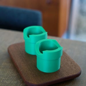 May include: Two bright green, cylindrical holders with a wide, flat rim and a distinct cutout section on the top edge. These containers feature a smooth, matte finish and are positioned together on a small, rectangular wooden base with rounded corners.