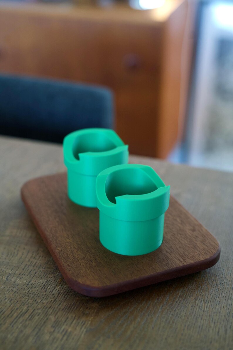 May include: Two bright green, cylindrical holders with a wide, flat rim and a distinct cutout section on the top edge. These containers feature a smooth, matte finish and are positioned together on a small, rectangular wooden base with rounded corners.