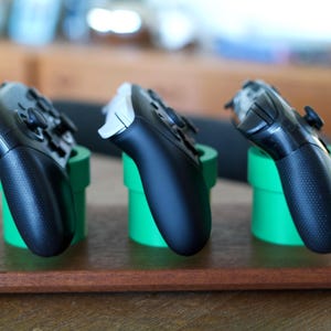 May include: Three video game controllers resting in three bright green cylindrical stands. The stands are mounted on a rectangular wooden base. Two of the controllers are black, and one controller is white and black, all angled upward.