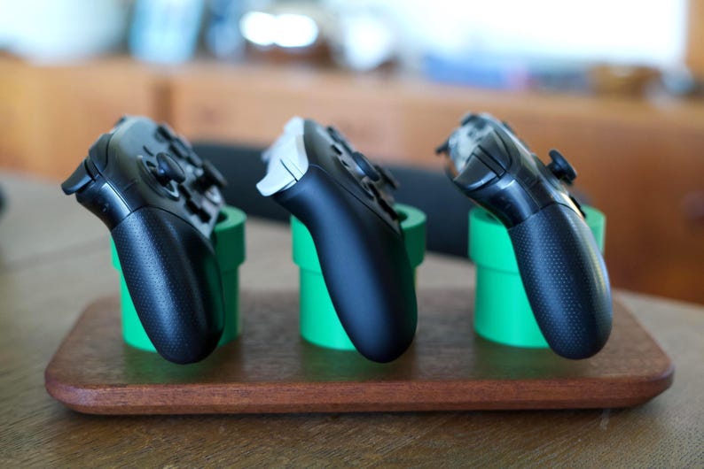 May include: Three video game controllers resting in three bright green cylindrical stands. The stands are mounted on a rectangular wooden base. Two of the controllers are black, and one controller is white and black, all angled upward.