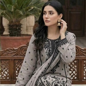 Puede incluir: Conjunto salwar kameez bordado gris claro. El atuendo incluye una túnica de manga larga, pantalones a juego y una bufanda drapeada, todos adornados con bordados florales negros. La mujer lleva pendientes dorados.
