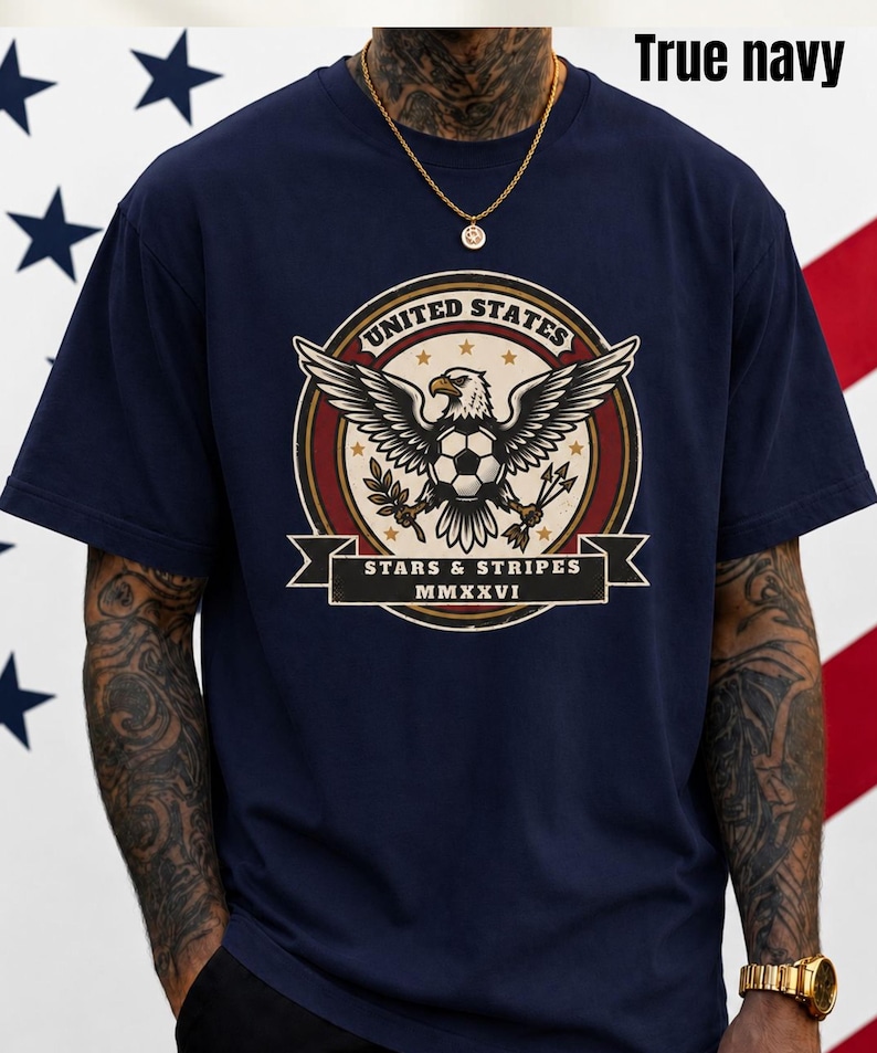 May include: A navy blue t-shirt featuring a graphic of an eagle with a soccer ball, wings spread, and the text "UNITED STATES" and "STARS & STRIPES". The text "MMXXVI" is also included. The shirt is labeled "True navy".