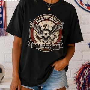 May include: Black t-shirt featuring a graphic of a bald eagle with outstretched wings, a soccer ball, and the text "UNITED STATES" and "STARS & STRIPES MMXXVI". The shirt is worn by a person with their hand in their pocket.