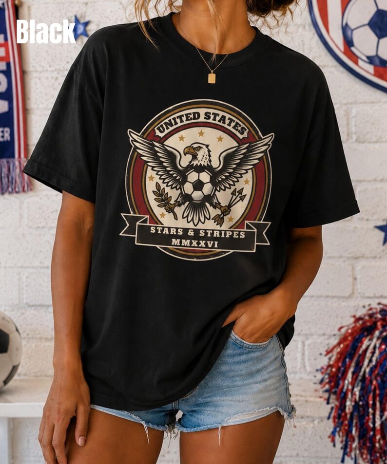 May include: Black t-shirt featuring a graphic of a bald eagle with outstretched wings, a soccer ball, and the text "UNITED STATES" and "STARS & STRIPES MMXXVI". The shirt is worn by a person with their hand in their pocket.