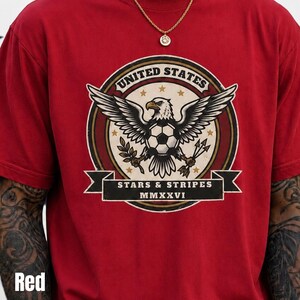 May include: Red t-shirt featuring a circular graphic with an eagle holding a soccer ball, olive branches, and arrows. The graphic includes the text "UNITED STATES" and "STARS & STRIPES MMXXVI".