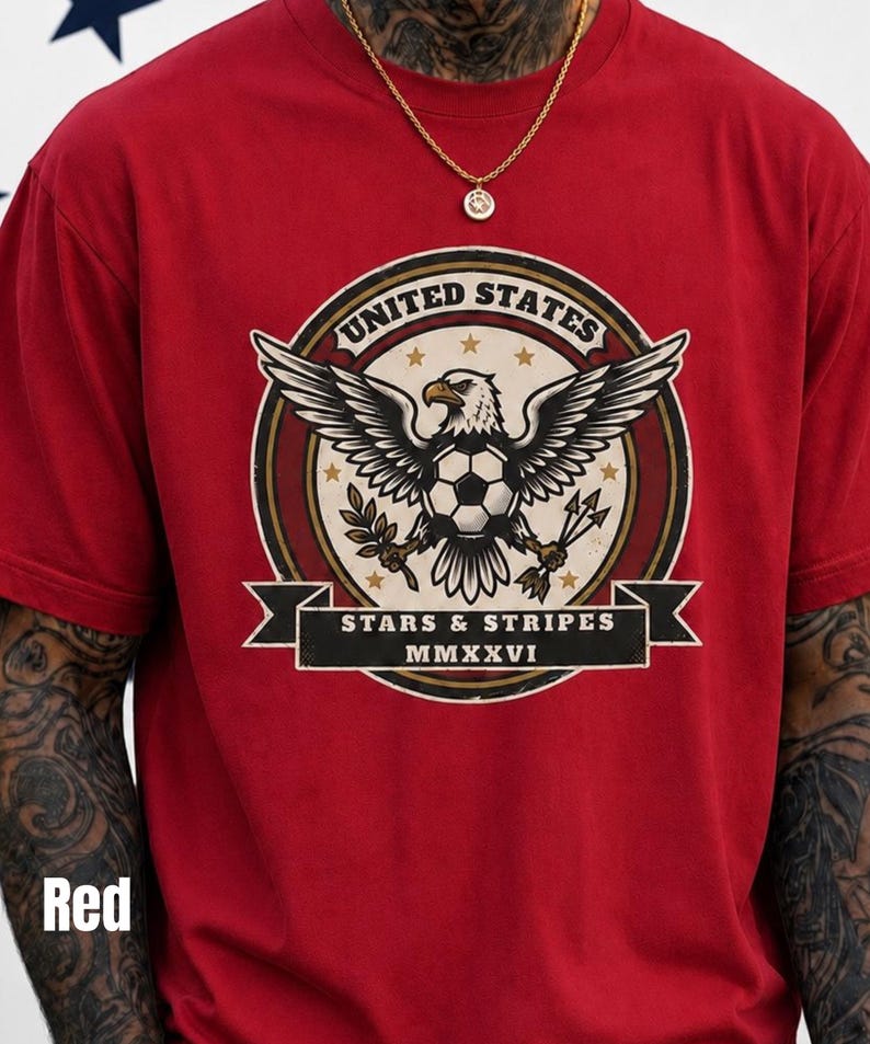 May include: Red t-shirt featuring a circular graphic with an eagle holding a soccer ball, olive branches, and arrows. The graphic includes the text "UNITED STATES" and "STARS & STRIPES MMXXVI".