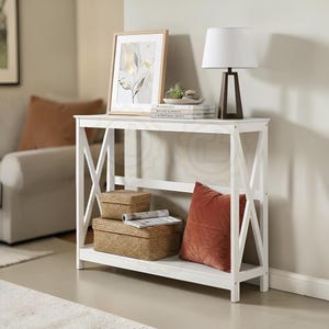 May include: A white console table with an X-frame design. The table is styled with a framed botanical print, a lamp with a white shade, and decorative items. Two woven storage boxes and a rust-coloured cushion are on the lower shelf.