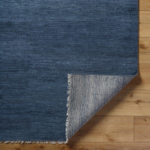 May include: A rectangular area rug featuring a deep navy blue woven surface with a visible texture. One corner of the rug is folded back to reveal a light gray, tightly woven backing material and a short, frayed fringe along the edge.