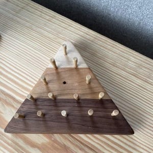 May include: A triangular wooden peg board with three sections of varying wood tones: light, medium, and dark brown. Small wooden pegs are inserted into the board. The board is on a light wood surface.