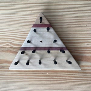 May include: A wooden triangle peg board with black pegs. The board has a light wood grain with a dark red stripe. The pegs are evenly spaced and arranged in a pattern. The board is on a light wood surface.