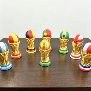 Op de afbeelding: Een verzameling miniatuur gouden trofeeën, elk bekroond met een bal beschilderd in de kleuren van verschillende nationale vlaggen. De trofeeën zijn gerangschikt op een donkerbruin oppervlak en tonen een verscheidenheid aan levendige kleuren.