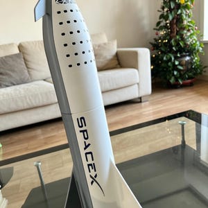 以下が含まれることがあります： 白いとグレーのSpaceX Starshipモデル。側面にSpaceXのロゴが印刷されています。モデルは、尖ったノーズ、フィン、黒いベースを備えた詳細なデザインです。モデルはガラスのテーブルに展示されています。
