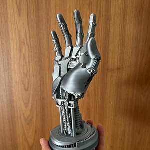 Puede incluir: Una escultura de mano robótica de color plateado con intrincados detalles mecánicos. La mano está en posición abierta, revelando la estructura interna. Está montada sobre una base circular. La escultura se sostiene en una mano.