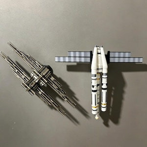 Könnte beinhalten: Zwei detaillierte Modellraumschiffe werden vor einem grauen Hintergrund gezeigt. Eines ist metallisch silber und schwarz, mehrzackig. Das andere ist eine weiß, schwarz und goldfarbene raketenartige Struktur mit einer gestreiften grau-schwarzen Plattform.