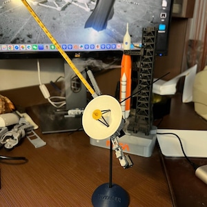 May include: A detailed model of the Voyager spacecraft, featuring a white dish antenna and various scientific instruments. The model is mounted on a blue base with the word "VOYAGER" printed on it. A yellow measuring stick extends from the spacecraft. Other space-themed models are in the background.