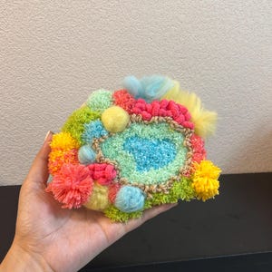 Può includere: Un'opera d'arte tessile colorata e strutturata con una forma centrale turchese circondata da accenti verdi, rosa, gialli e arancioni. Il pezzo è adornato con pompon e texture soffici, creando un'estetica vibrante e fatta a mano.