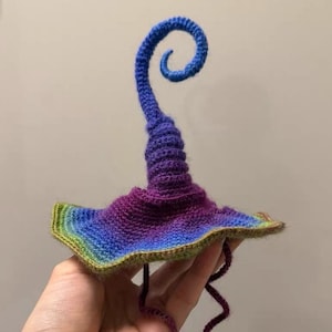 Pode incluir: Um chapéu de crochê artesanal em um arco-íris vibrante de cores. O chapéu apresenta uma aba alargada e um topo alto e em espiral. O gradiente de cores transita do verde para o azul, roxo e, finalmente, um azul escuro no topo. Duas mechas roxas pendem da aba.