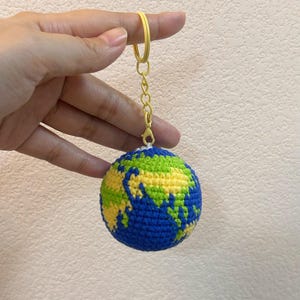 K&ouml;nnte beinhalten: Ein geh&auml;kelter Globus-Schl&uuml;sselanh&auml;nger mit blauen Ozeanen und gr&uuml;nen und gelben Kontinenten. Der Schl&uuml;sselanh&auml;nger hat einen gelben Ring und eine Kette. Der Globus wird von einer Hand vor einem neutralen Hintergrund gehalten.