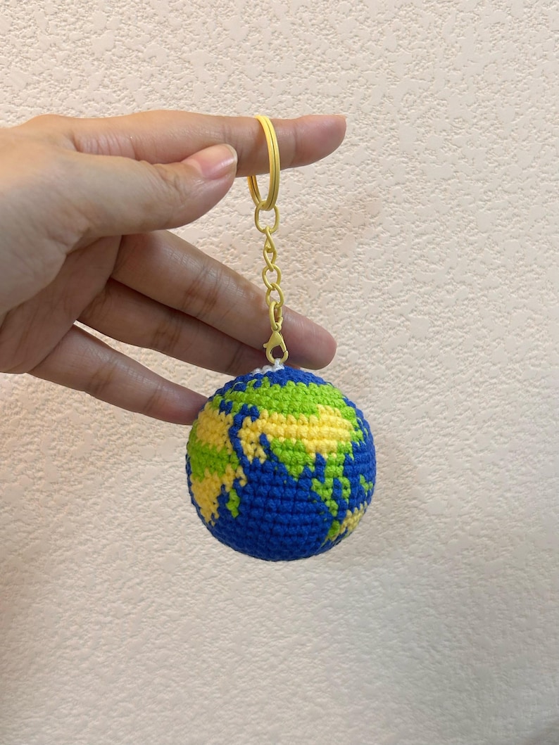 K&ouml;nnte beinhalten: Ein geh&auml;kelter Globus-Schl&uuml;sselanh&auml;nger mit blauen Ozeanen und gr&uuml;nen und gelben Kontinenten. Der Schl&uuml;sselanh&auml;nger hat einen gelben Ring und eine Kette. Der Globus wird von einer Hand vor einem neutralen Hintergrund gehalten.