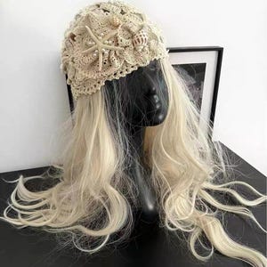 May include: A beige crocheted head covering adorned with seashells, pearls, and a starfish. The head covering is displayed on a black mannequin head with long, wavy, blonde hair. The head covering is a unique accessory.