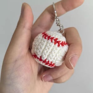 May include: A white crocheted baseball keychain with red stitching details. The baseball is round and attached to a silver keyring. The keychain is held in a hand.