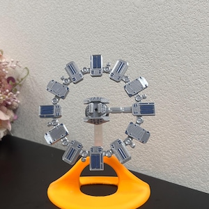 May include: A silver-coloured model of a rotating space station featuring a central hub connected by a single arm to a circular ring of twelve rectangular modules with blue accents, all mounted on a bright orange curved plastic base stand.