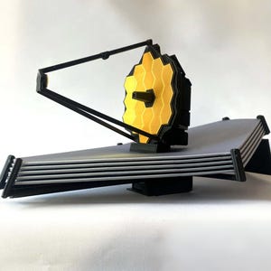 May include: A 3D-printed model of the James Webb Space Telescope. The model features a gold-colored, hexagonal primary mirror, a black support structure, and a silver-colored sunshield. The model is displayed on a white background.