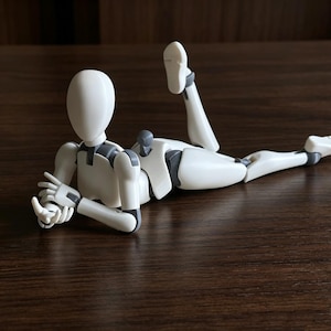 Op de afbeelding: Een witte, gelede figuur met grijze gewrichten en accenten, liggend op een donkerbruin houten oppervlak. De figuur ligt in een liggende houding, met één been uitgestrekt en het andere gebogen. De armen zijn gebogen en de handen open.