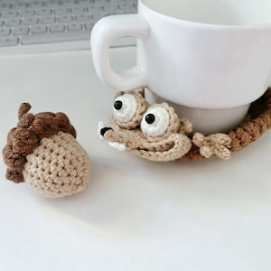 Puede incluir: Juego de posavasos de bellota y ardilla de ganchillo. La bellota es de color beige con una tapa marrón. El posavasos de ardilla es de color beige con ojos blancos grandes y pupilas negras. Una taza blanca se encuentra sobre el posavasos de ardilla.