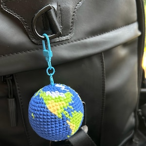 May include: A blue, green, and white crocheted globe keychain hanging from a black backpack. The globe features continents in green and yellow against a blue ocean. The keychain is attached with a light blue clasp.