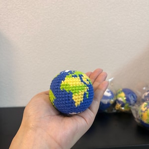 K&ouml;nnte beinhalten: Eine geh&auml;kelte Weltkugel in Blau, Gr&uuml;n und Gelb, gehalten in einer Hand. Die Weltkugel zeigt Kontinente in Gr&uuml;n und Gelb, mit blauen Ozeanen. Weitere Globusse sind im Hintergrund zu sehen.