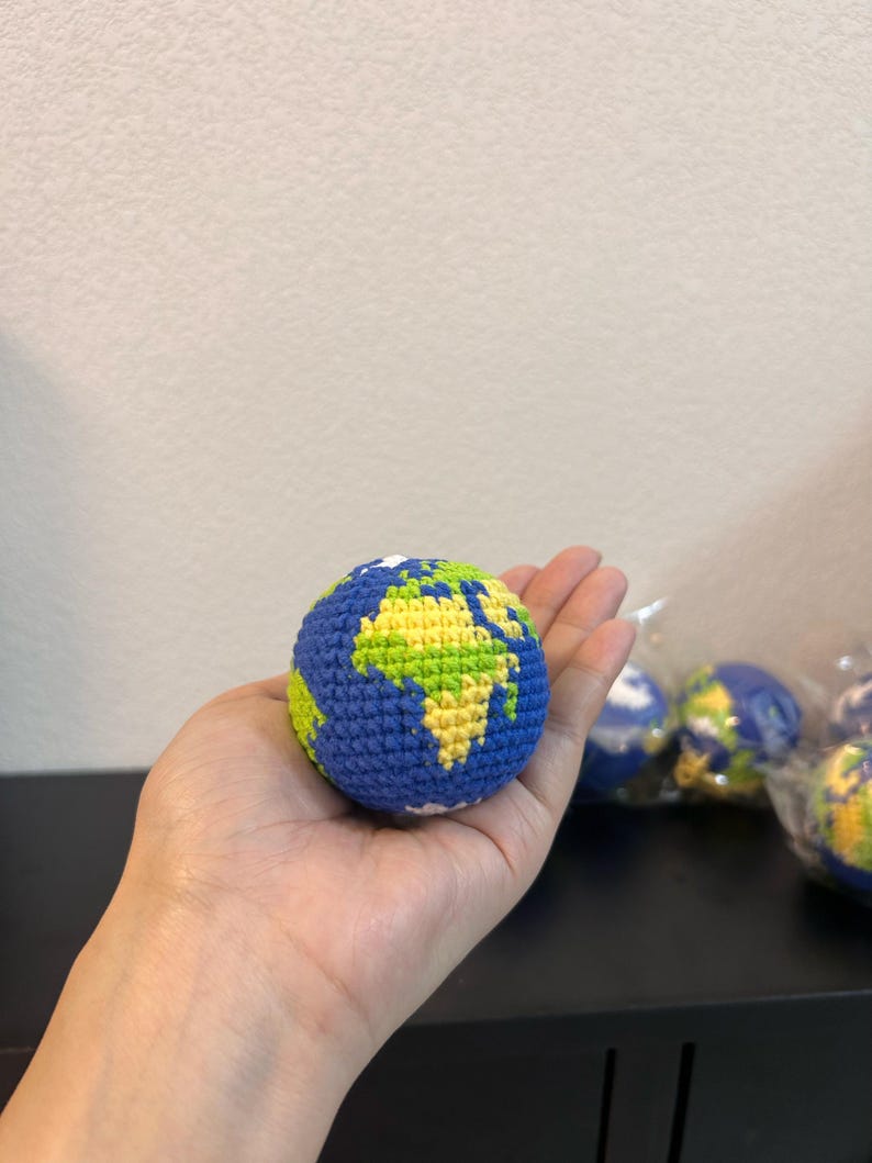K&ouml;nnte beinhalten: Eine geh&auml;kelte Weltkugel in Blau, Gr&uuml;n und Gelb, gehalten in einer Hand. Die Weltkugel zeigt Kontinente in Gr&uuml;n und Gelb, mit blauen Ozeanen. Weitere Globusse sind im Hintergrund zu sehen.