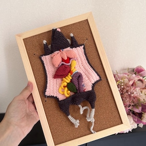 May include: A framed crochet artwork depicting a stylized anatomical figure. The piece features various colored crochet elements, including pink, red, yellow, and purple, against a light pink background. The frame is made of light-colored wood.