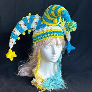 May include: A whimsical, handmade crochet hat in blue, yellow, and white, adorned with star and pom-pom accents. The jester-style hat features a long, pointed tip and is displayed on a mannequin head with colorful hair extensions. The hat is a unique, eye-catching accessory.