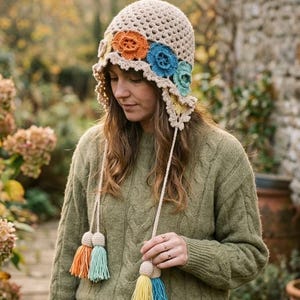 May include: A beige crochet hat with colorful floral accents and dangling tassels. The hat features orange, blue, and green flower designs along the brim. The tassels are in shades of orange, teal, yellow, and blue. The hat is a unique, handmade accessory.