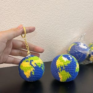 Puede incluir: Llavero de globo de ganchillo con océanos azules y continentes verdes y amarillos. El globo está unido a un llavero de color dorado. Se ven otros globos de ganchillo en el fondo.