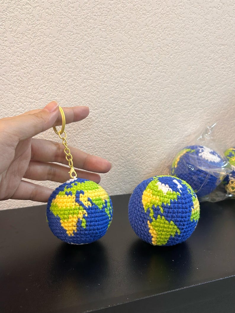 K&ouml;nnte beinhalten: Geh&auml;kelter Globus-Schl&uuml;sselanh&auml;nger mit blauen Ozeanen und gr&uuml;nen und gelben Kontinenten. Der Globus ist an einem goldfarbenen Schl&uuml;sselanh&auml;nger befestigt. Weitere geh&auml;kelte Globen sind im Hintergrund zu sehen.