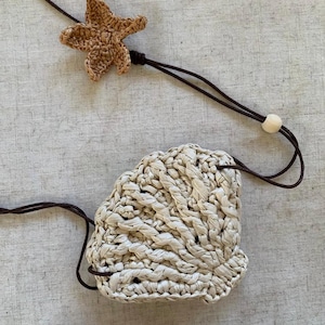 Può includere: Un astuccio intrecciato bianco sporco con chiusura a coulisse e un ciondolo a forma di stella marina. La stella marina e l'astuccio sono realizzati in un materiale di fibra naturale. Il cordoncino è marrone scuro e c'è una perlina di legno sul cordoncino.