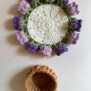 May include: A handmade crochet doily with a floral design. The doily features a white center, a green border, and purple and lavender flower accents. Below is a small, brown crochet basket.