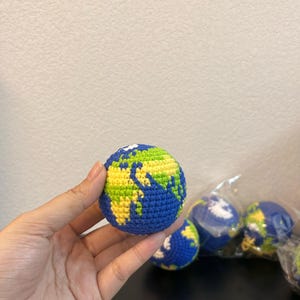 K&ouml;nnte beinhalten: Ein geh&auml;kelter Globus in Blau, Gr&uuml;n und Gelb. Der Globus wird in einer Hand gehalten, mit weiteren &auml;hnlichen Globussen im Hintergrund. Die Kontinente sind in Gr&uuml;n und Gelb dargestellt, mit blauen Ozeanen.