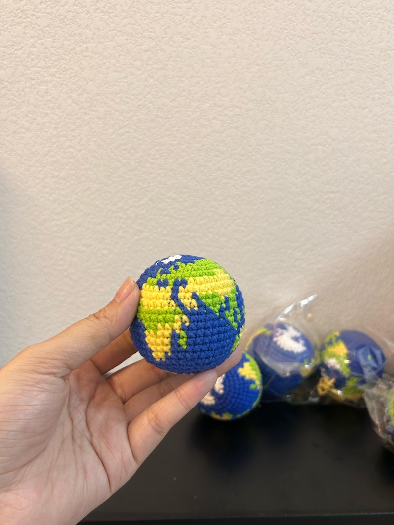 K&ouml;nnte beinhalten: Ein geh&auml;kelter Globus in Blau, Gr&uuml;n und Gelb. Der Globus wird in einer Hand gehalten, mit weiteren &auml;hnlichen Globussen im Hintergrund. Die Kontinente sind in Gr&uuml;n und Gelb dargestellt, mit blauen Ozeanen.
