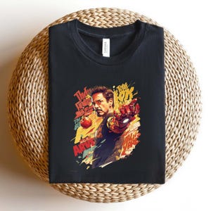 May include: Black t-shirt featuring a colorful graphic of a man with a red and gold mechanical arm, with text that reads "The New Avengers" and "Tony Stark". The shirt is folded on a woven surface.