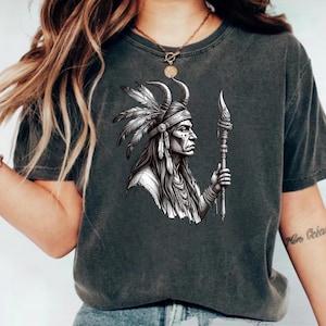 May include: Dark gray t-shirt featuring a detailed black and white illustration of a Native American figure in profile, wearing a feathered headdress and holding a staff. The design is centered on the shirt.